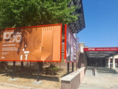 Oranžová reklamní tabule u chodníku před budovou, vedle billboardu a zelených stromů, jasná obloha.
