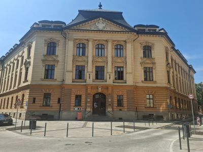 Historická kamenná budova s centrálním štítem, sloupy a řadou oken, za jasného modrého nebe.