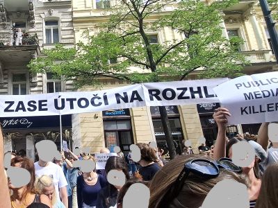 Dav lidí drží transparent s nápisem „ZASE ÚTOČÍ NA ROZHLAS“ před budovou v ulici.