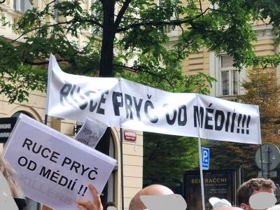 Protestní transparenty s nápisy „Ruce pryč od médií“ a „Prvč do médií“ před budovou, dav lidí.