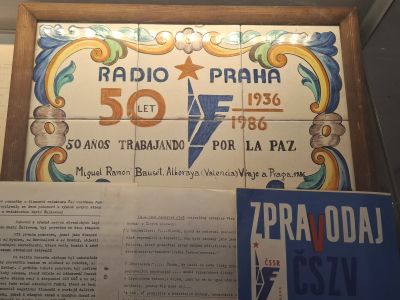 Dřevěný plakát s nápisem RADIO PRAHA, letopočty a novinové výstřižky pod ním.