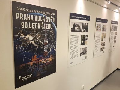 Výstavní plakát „Praha volá svět!“ a další informační panely na stěně galerie, osvětlené reflektory.