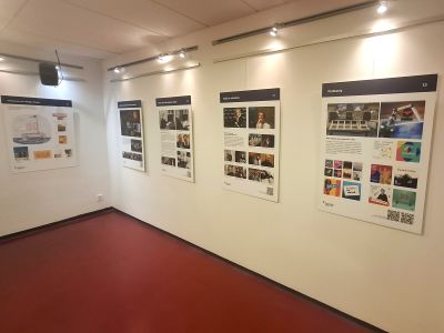 Galerie s několika nástěnnými panely a fotografiemi, osvětlená stropními světly, prázdná výstavní místnost.