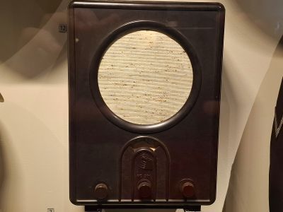 Hnědý starožitný rádiový přijímač s kruhovým reproduktorem a ladicím knoflíkem na přední straně.