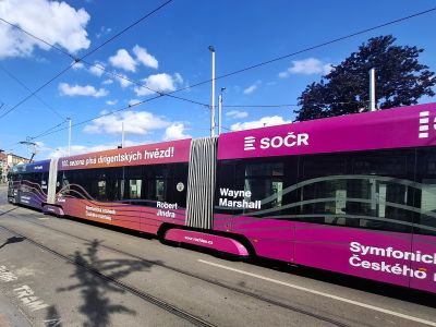 Růžový tramvajový vůz s reklamou a nápisem SOČR projíždí ulicí pod modrou oblohou.